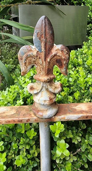 Rusted fleur-de-lis fence finial over green shrubs